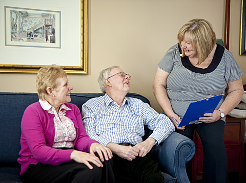 Photo of a mature couple sitting on a sofa and smiling toward a staff member with a clipboard, reflecting supportive care in residential settings